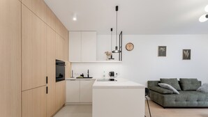 Een moderne keuken met houten kastjes, een wit aanrecht en een inbouwoven. In de woonruimte staat een groene bank met kussens.
