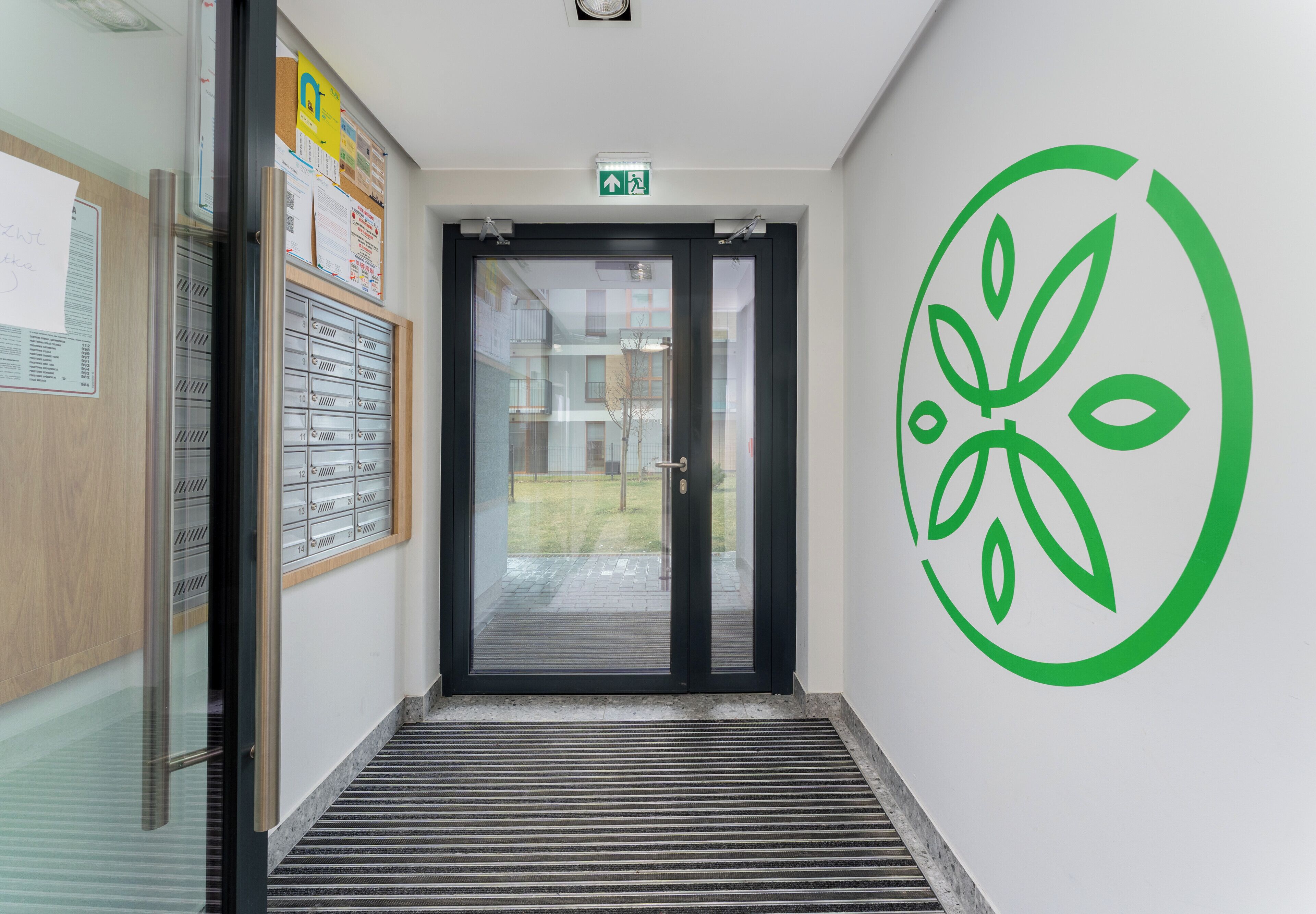 Een hal met een glazen deur die naar buiten leidt, een groen logo op de muur en een prikbord met papieren.