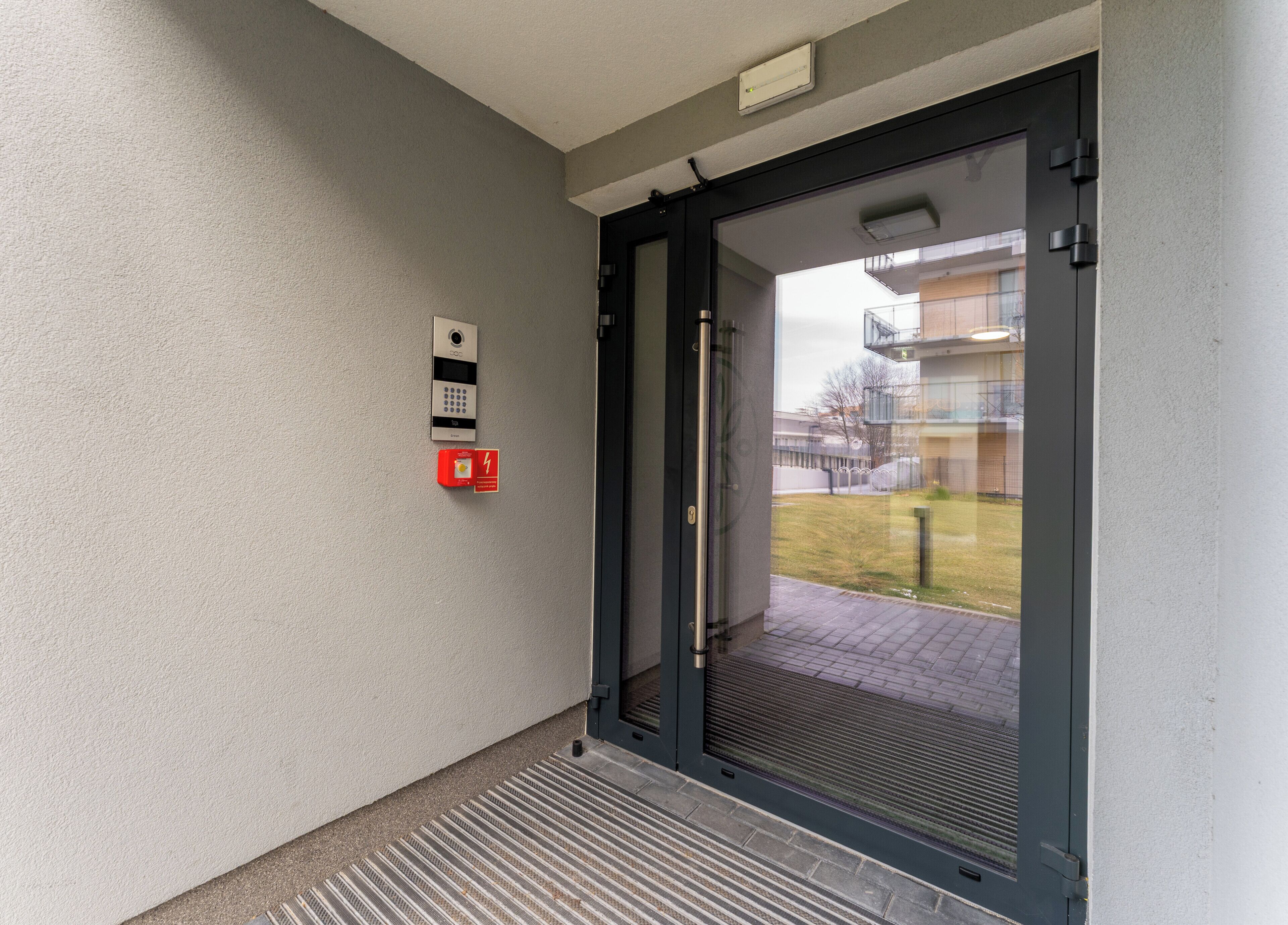 Een moderne entree met glazen deur, brandmelder en een intercom.