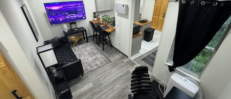 Un espace de vie compact comprenant un coin repas, un téléviseur fixé au mur, une petite cuisine et une fenêtre avec des rideaux.