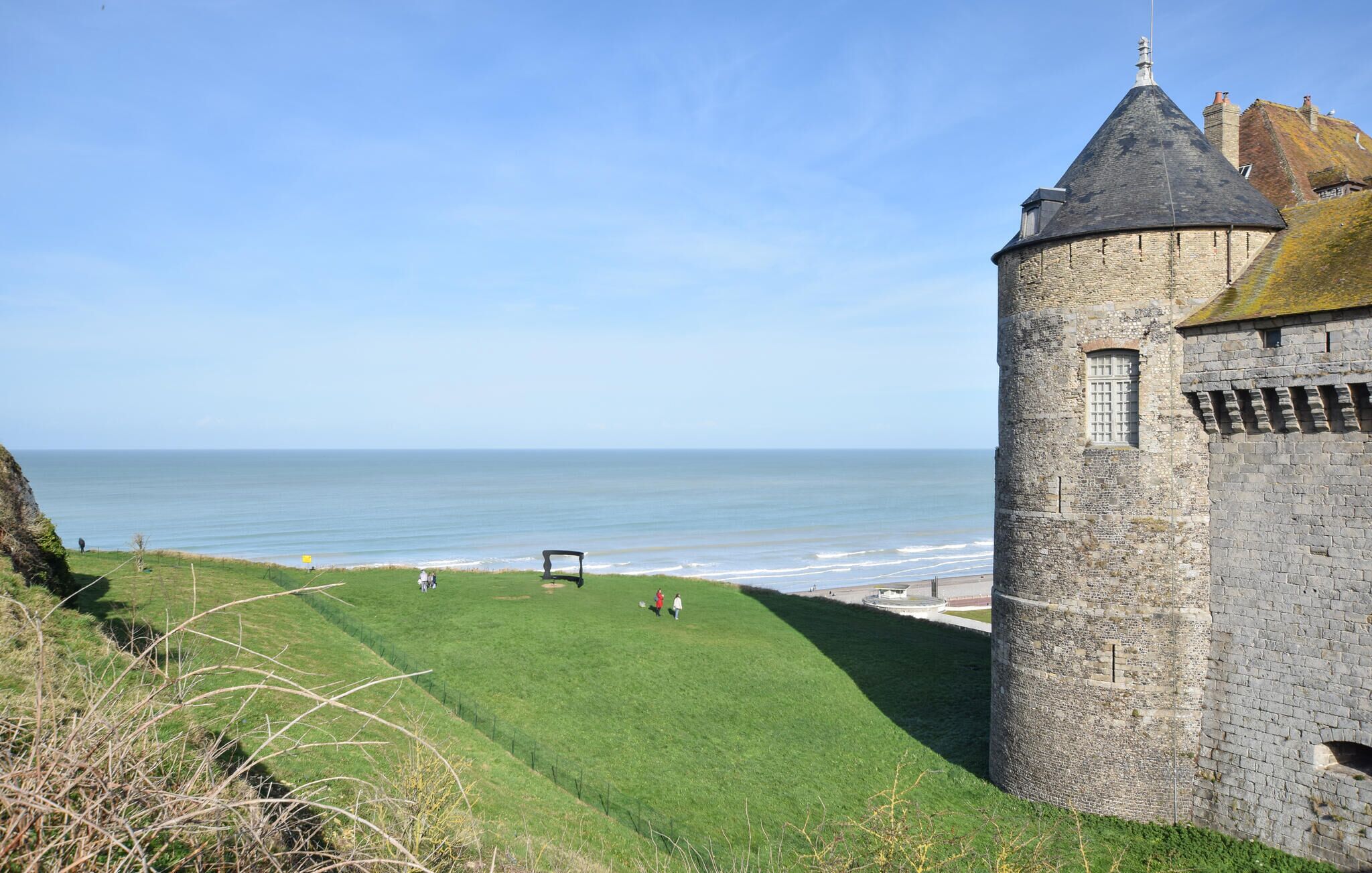 Uma torre de pedra com telhado cónico, com vista para uma área gramada e o mar.