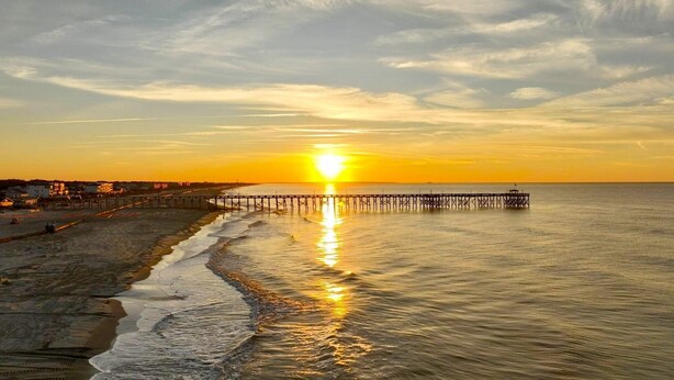 Une jetée s’avançant dans des eaux calmes au coucher du soleil, avec une plage sur la gauche.
