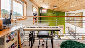 Una habitación con techo de madera, pared verde, un piano, un escritorio con computadora y ventana con vista a árboles.