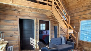 Interior acogedor de una cabaña con cama en un altillo, sofá, sillón, mesita y escalera.