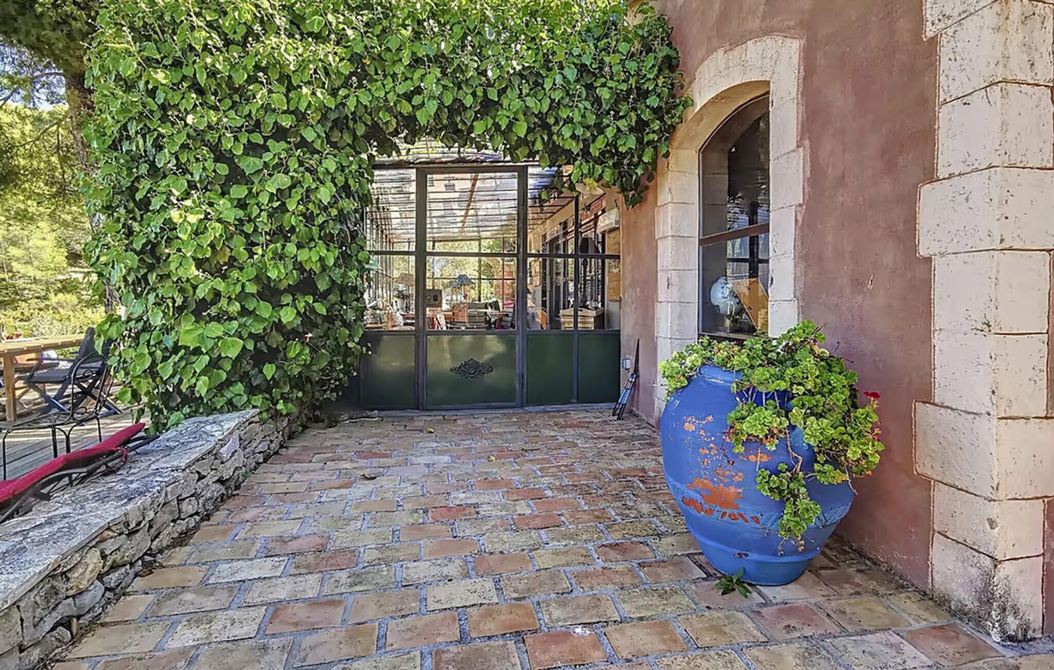 A stone-paved courtyard with a glass door, a large blue planter with plants, and a brick wall.