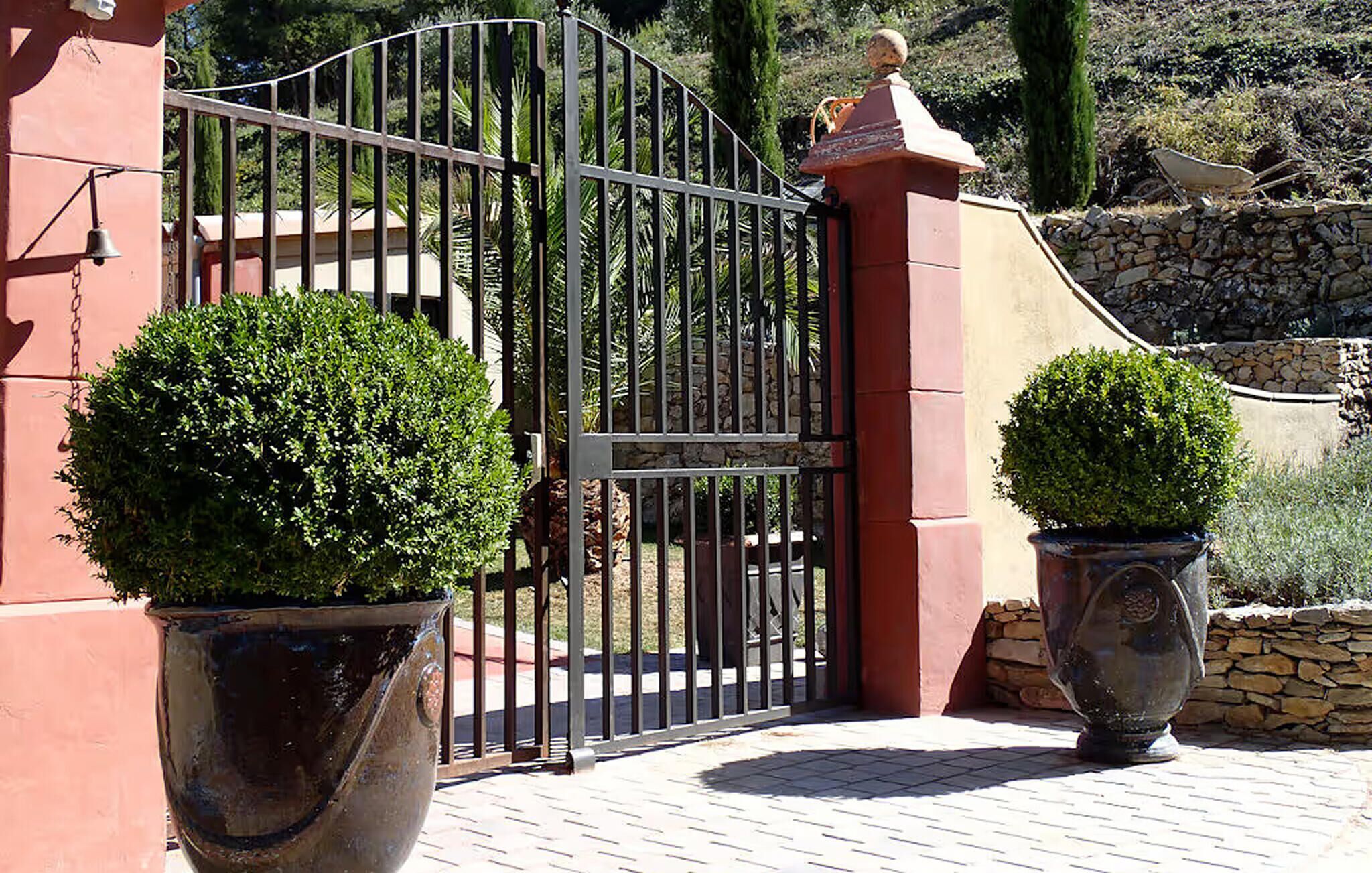 Uma entrada murada com portão de metal, plantas em vasos e parede de pedra.