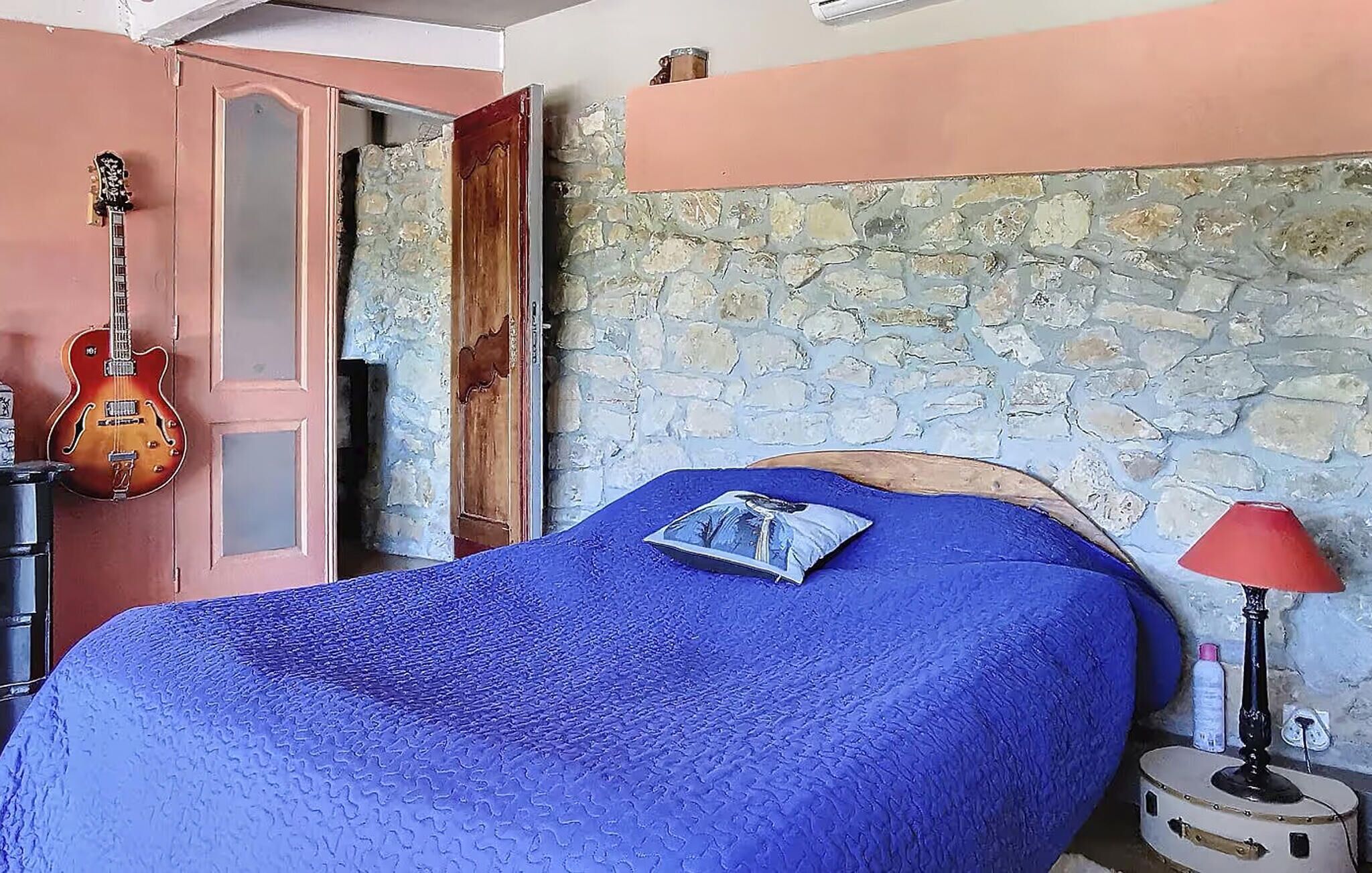 A bedroom with a stone wall, a bed with a blue blanket, a guitar, and a red lamp.