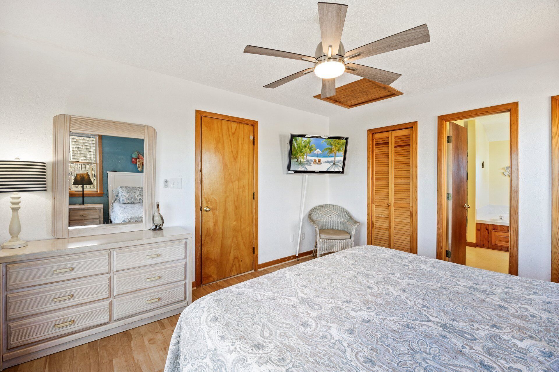 A bedroom with a bed, dresser, vanity, mirror, ceiling fan, TV, and wooden doors.
