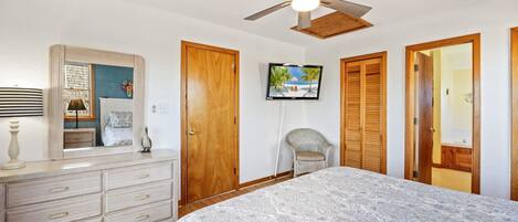 A bedroom with a bed, dresser, vanity, mirror, ceiling fan, TV, and wooden doors.