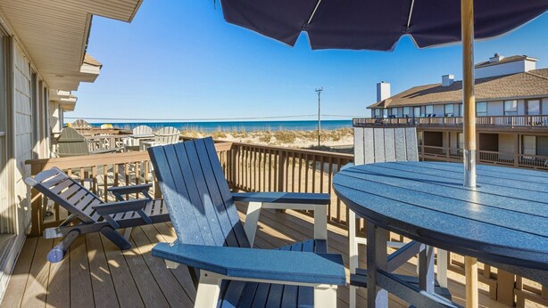 Een houten terras met blauwe Adirondack-stoelen en een ronde tafel, met uitzicht op een strand onder een helderblauwe lucht.