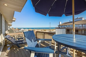 A wooden deck with blue Adirondack chairs and a round table, overlooking a beach with a clear blue sky. - Summer Breeze w/ Ocean views Comm pool/tennis (Kitty Hawk)