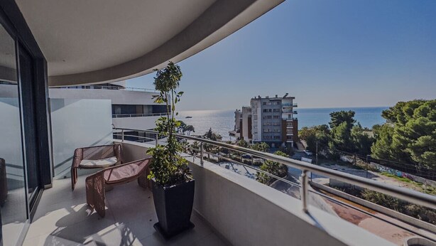 Een balkon met rieten meubilair en een bloempot, met uitzicht op een kustgebied met gebouwen en een heldere lucht.