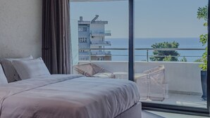 Een slaapkamer met een groot bed, een balkon met rieten meubilair en uitzicht op de zee en gebouwen.