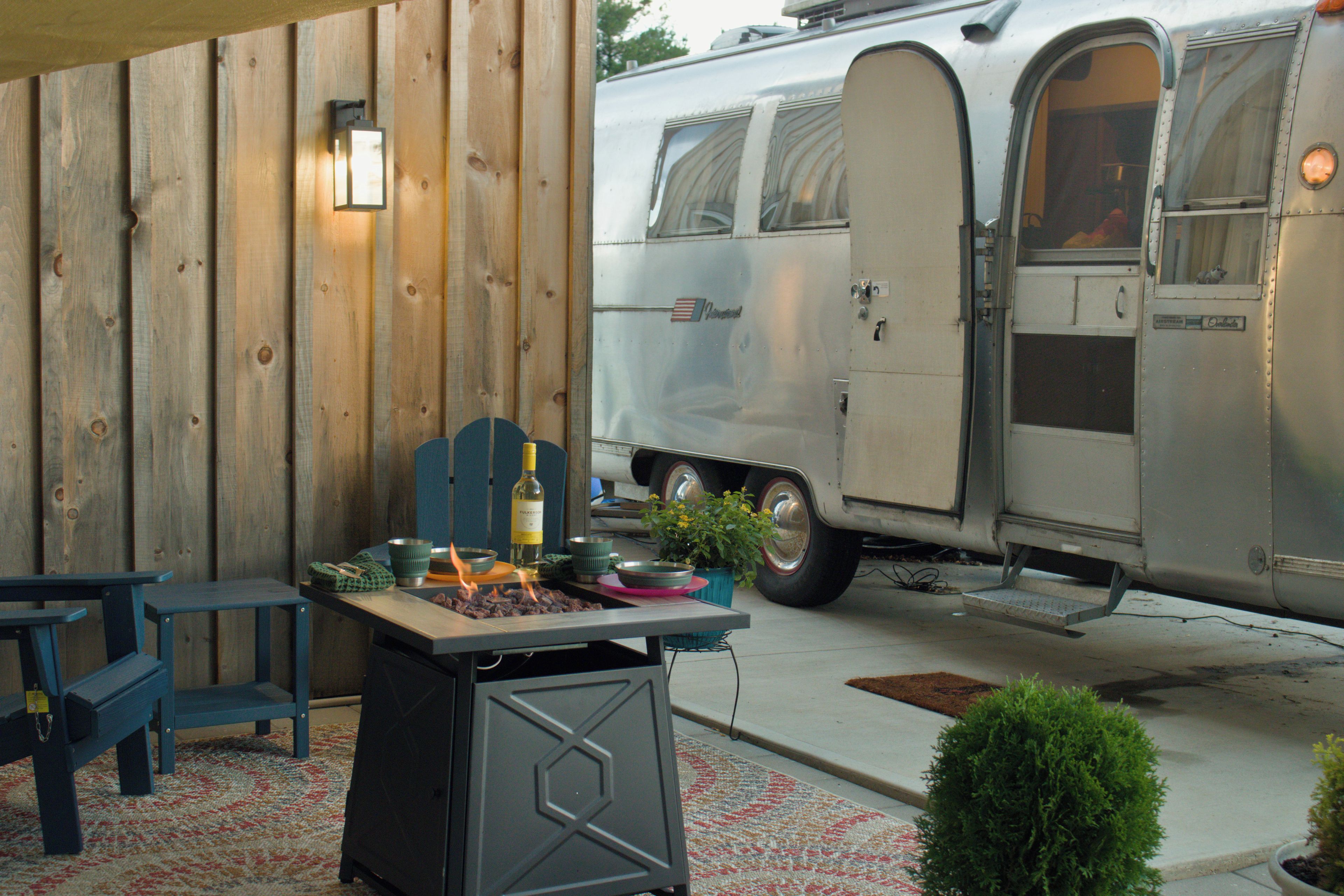 A vintage camper van parked next to a wooden wall with a table set for a meal.