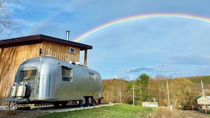 Een oude caravan met een houten blokhut ernaast, geparkeerd op een grasveld met een regenboog op de achtergrond.