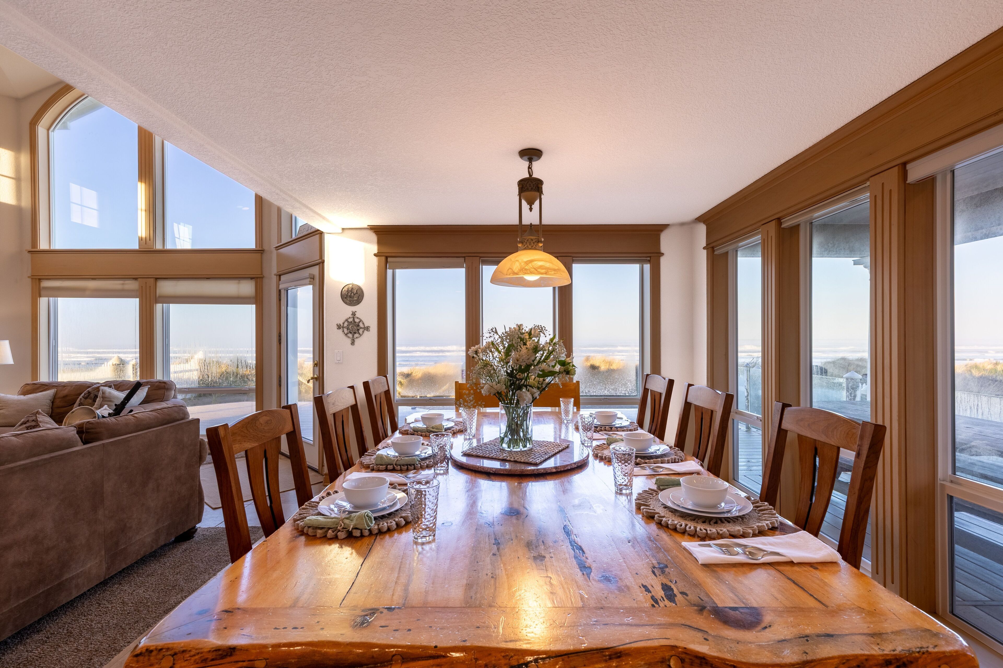 A dining room with a large wooden table set for six, surrounded by chairs, with a vase of flowers in the center.