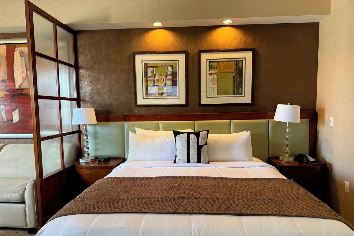 Une chambre d’hôtel avec un grand lit, deux tables de chevet avec des lampes, une tête de lit et deux œuvres d’art encadrées accrochées au mur.