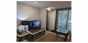 A living room with a flat-screen TV, a lamp, a small table, and a window with curtains.