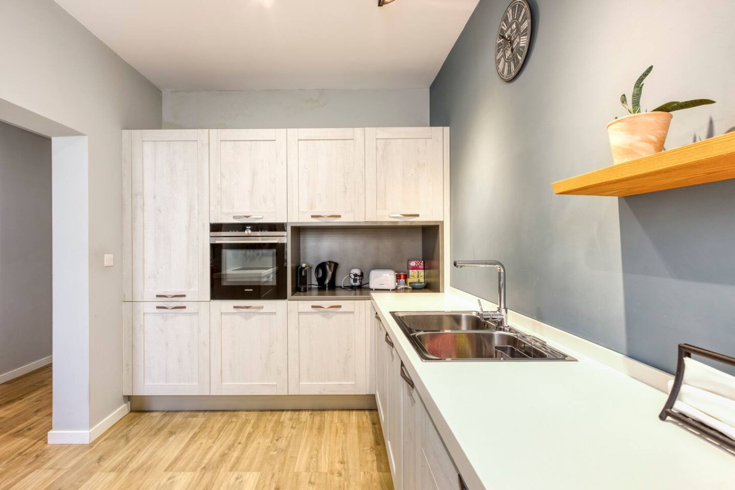 Une cuisine moderne avec des meubles de couleur bois clair, un évier en acier inoxydable et une étagère en bois sur laquelle est posée une plante en pot.