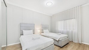 Habitación de hotel con dos camas, una mesita de noche con lámpara, una ventana con cortinas translúcidas y suelo de madera.