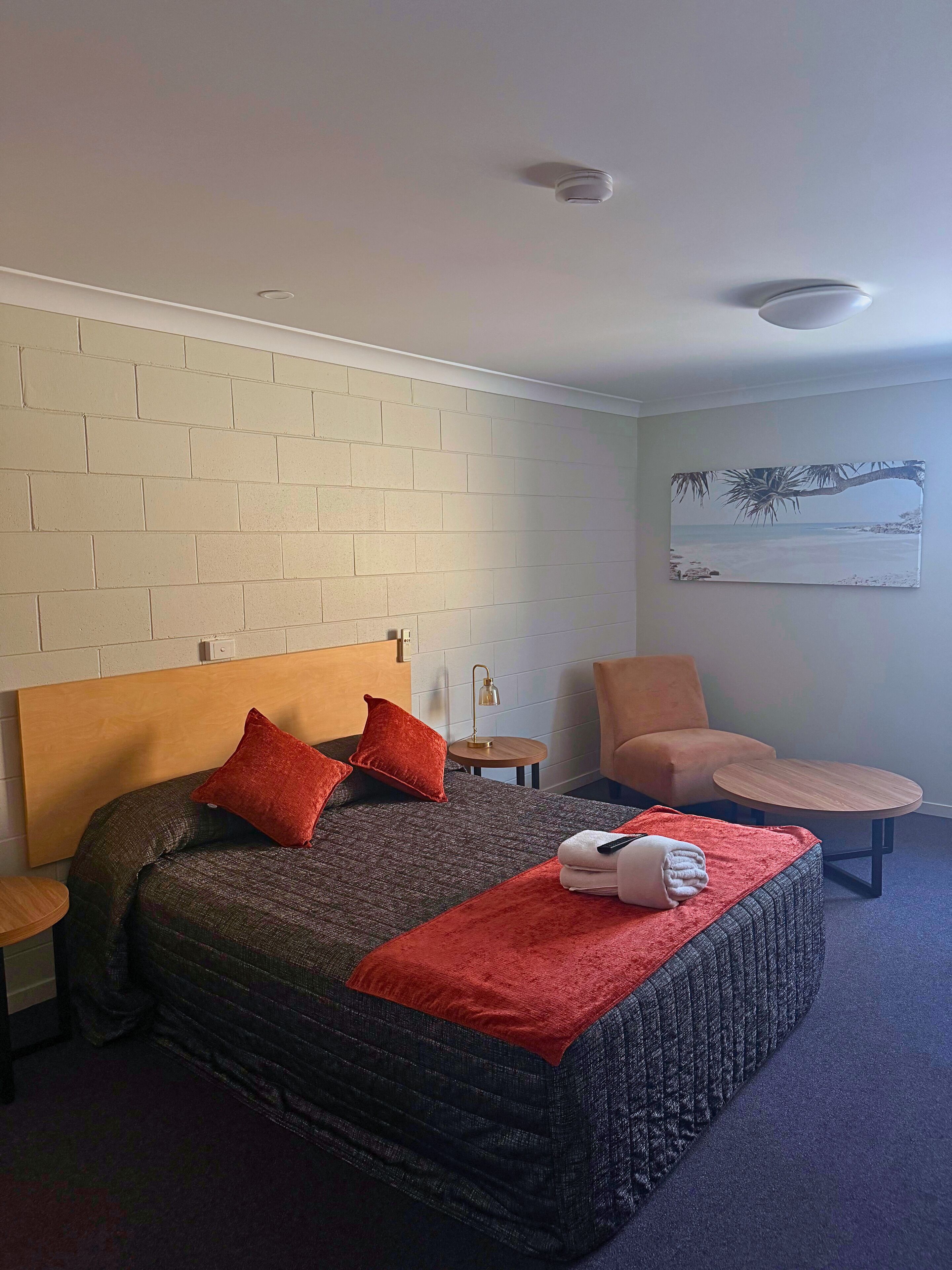 A hotel room with a bed, a chair, a small table, a painting on the wall, and a brick wall.