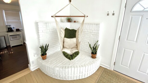 A room with a white brick wall, a white door, and a hanging chair with cushions. There are two potted plants on either side of the chair. - Peaceful + Cozy 2BR Oasis (Dayton)