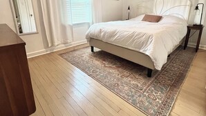 A bedroom with a bed, a nightstand, a lamp, a window with curtains, a door, and a rug. - Peaceful + Cozy 2BR Oasis (Dayton)
