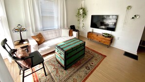 A modern living room with a white sofa, a colorful ottoman, a flat-screen TV, and a wooden cabinet. - Peaceful + Cozy 2BR Oasis (Dayton)