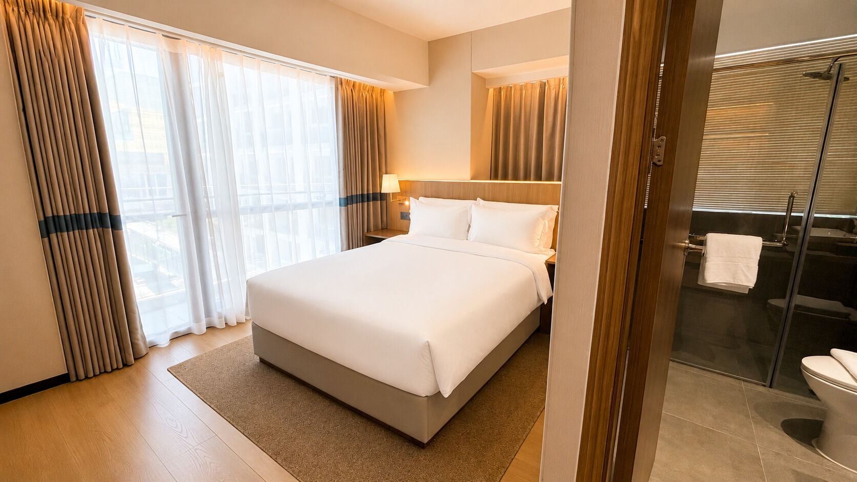 A hotel room with a large bed, a bedside table, a lamp, a bathroom with a shower, and a glass door.