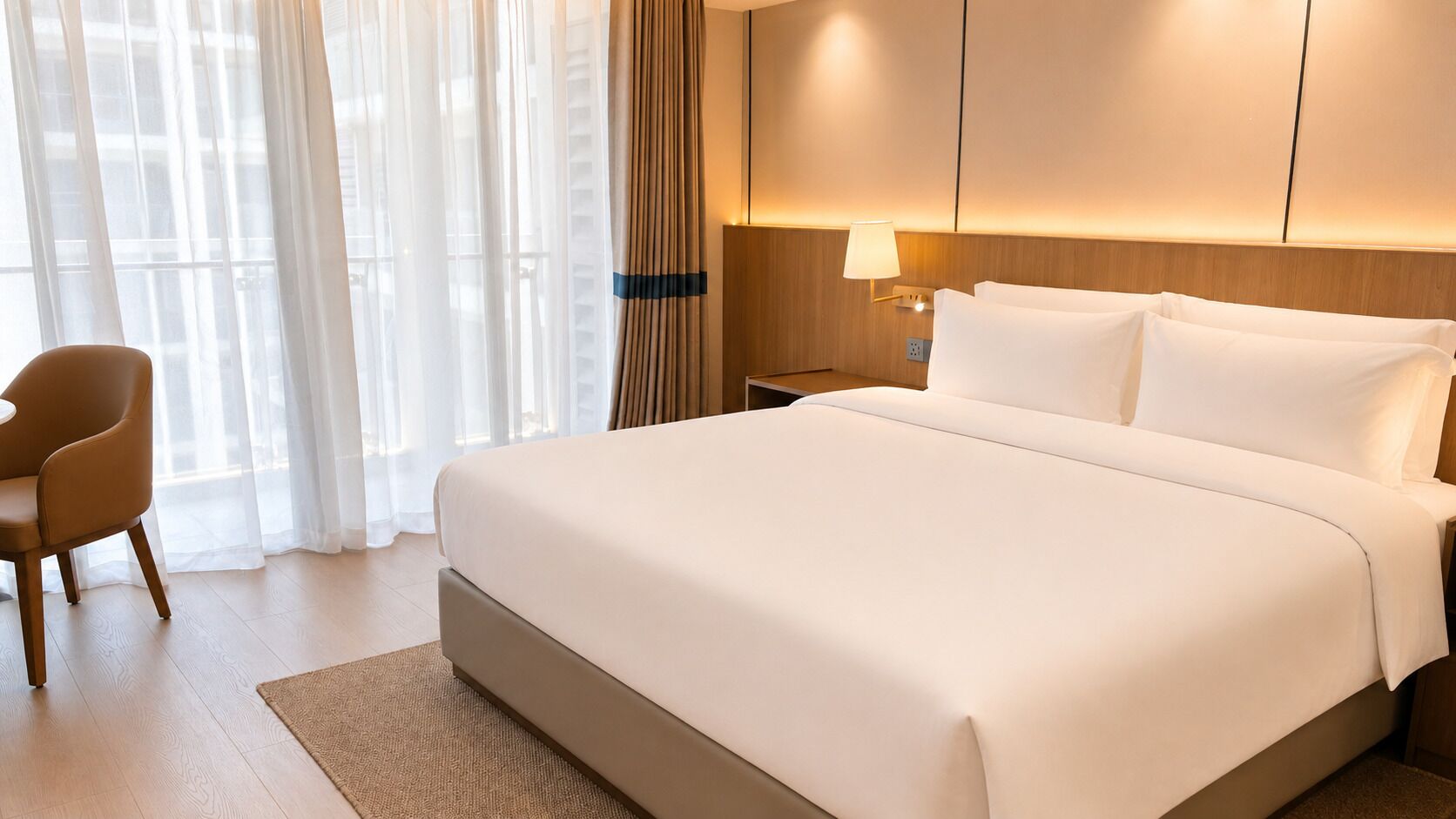 Ein Hotelzimmer mit einem großen Bett, einem Nachttisch mit Lampe, einem Sessel und durchsichtigen Vorhängen.