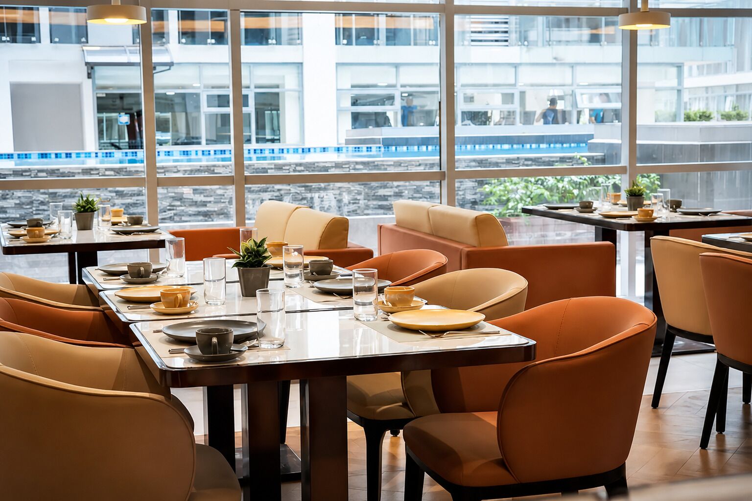 Een moderne eetruimte met tafels voor twee personen, grote ramen met uitzicht op een gebouw en een trein, en oranje stoelen.