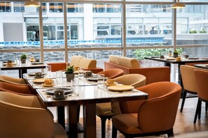 A modern dining area with tables set for two, large windows with a view of a building and a train, and orange chairs. - Wyndham Garden Manila Bay (Parañaque)