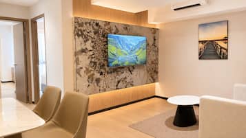 Um saguão de hotel moderno com uma grande TV de tela plana montada em uma parede texturizada, uma área de refeições com mesa de mármore e cadeiras, e um espaço de estar acolhedor com uma pequena mesa redonda.