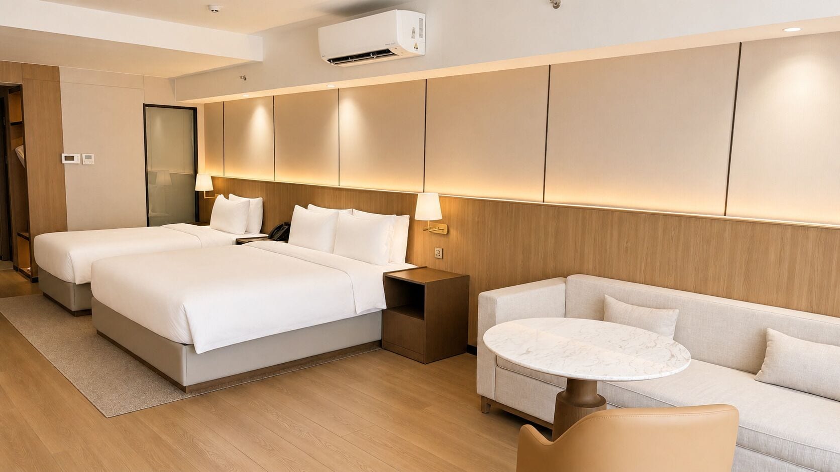 A hotel room with a large bed, a sofa, a small round table, and a wall-mounted air conditioner.