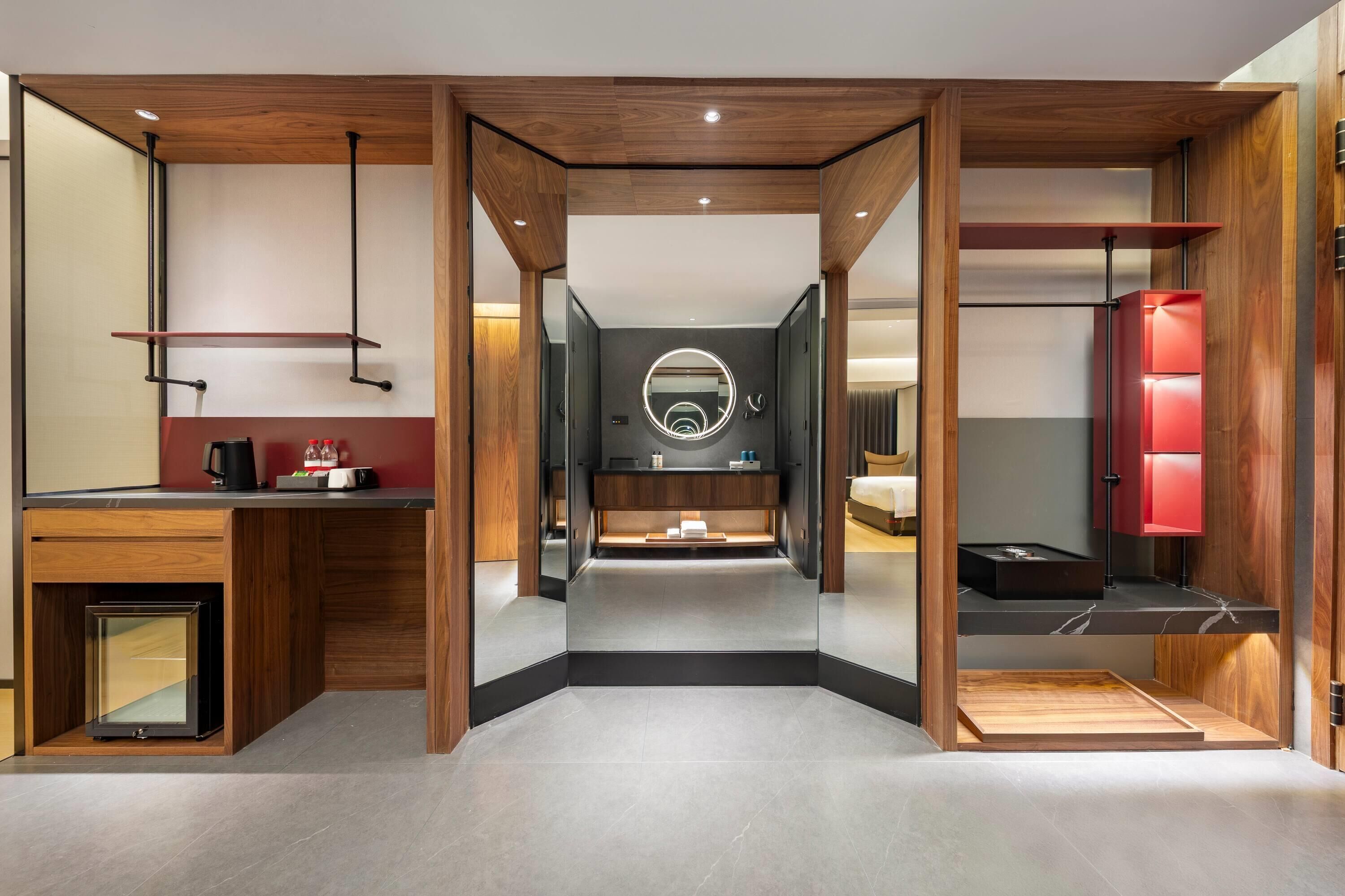 Una habitación de hotel moderna con un espejo central, un tocador con un espejo redondo, una cama y un armario rojo.