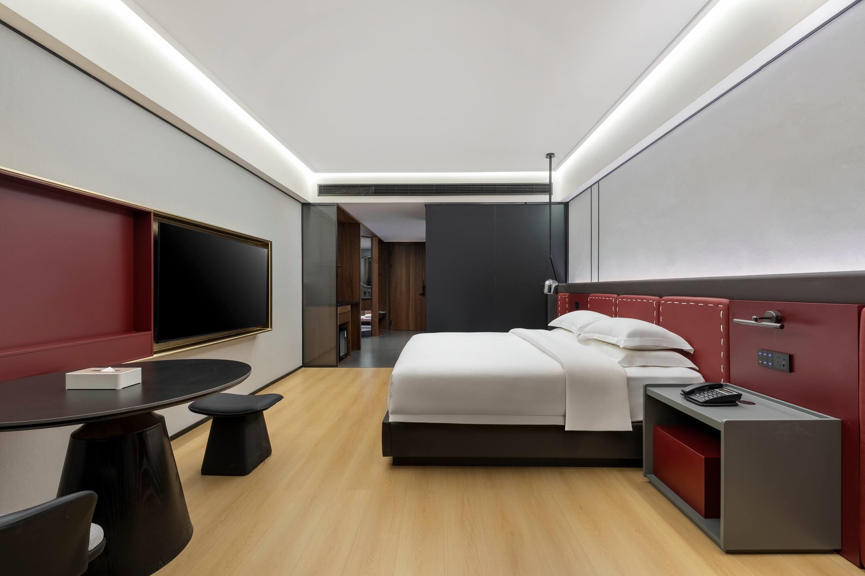 A modern hotel room with a bed, bedside table, TV, and a desk with a chair.