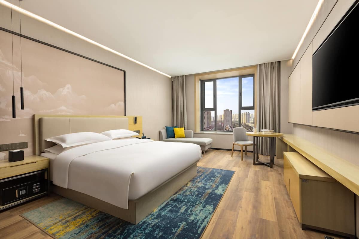 A modern hotel room with a large bed, a desk, a chair, a TV, and a city view.