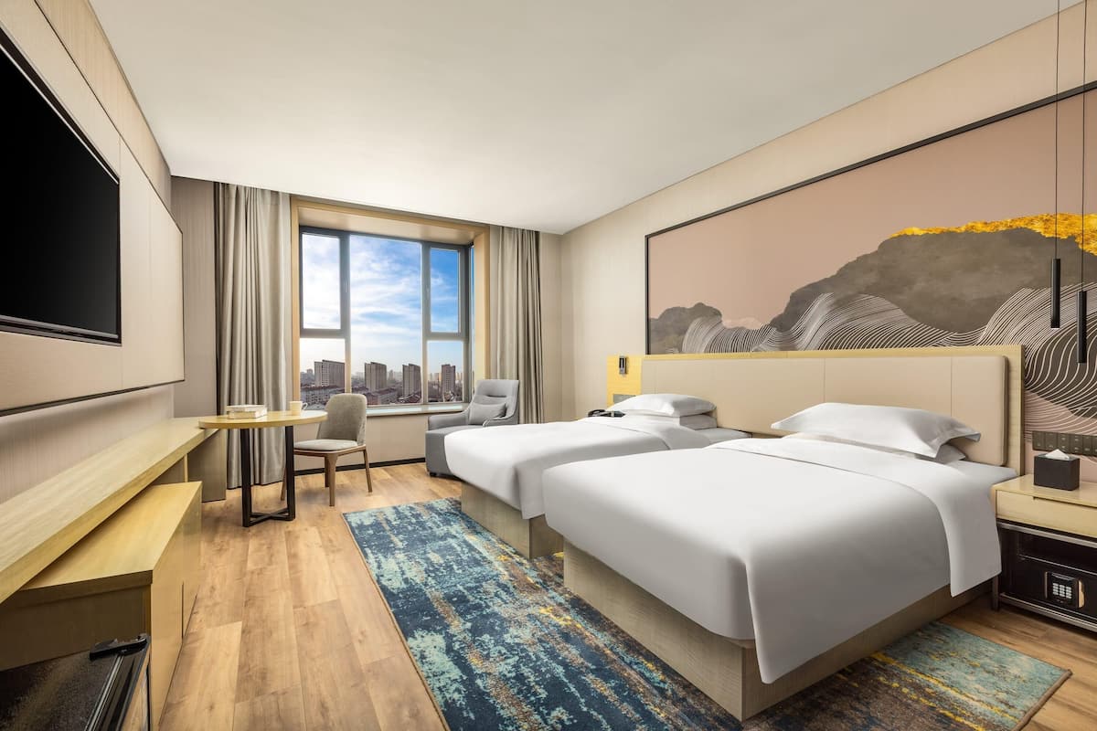 A hotel room with two beds, a desk, a chair, a TV, and a large window with a city view.