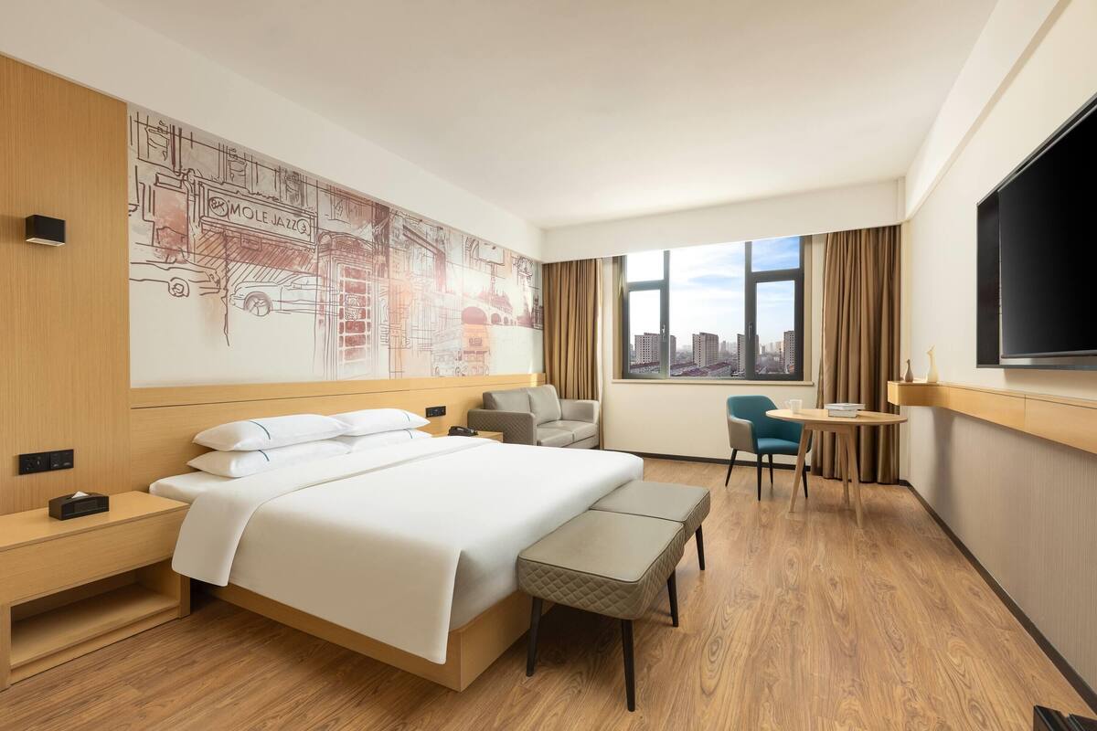 A hotel room with a large bed, a desk, a chair, a TV, and a city view.