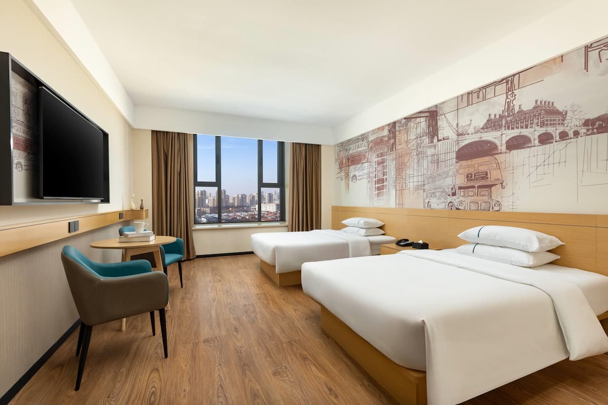 A hotel room with a bed, a desk, two chairs, a TV, and a large window with a city view.