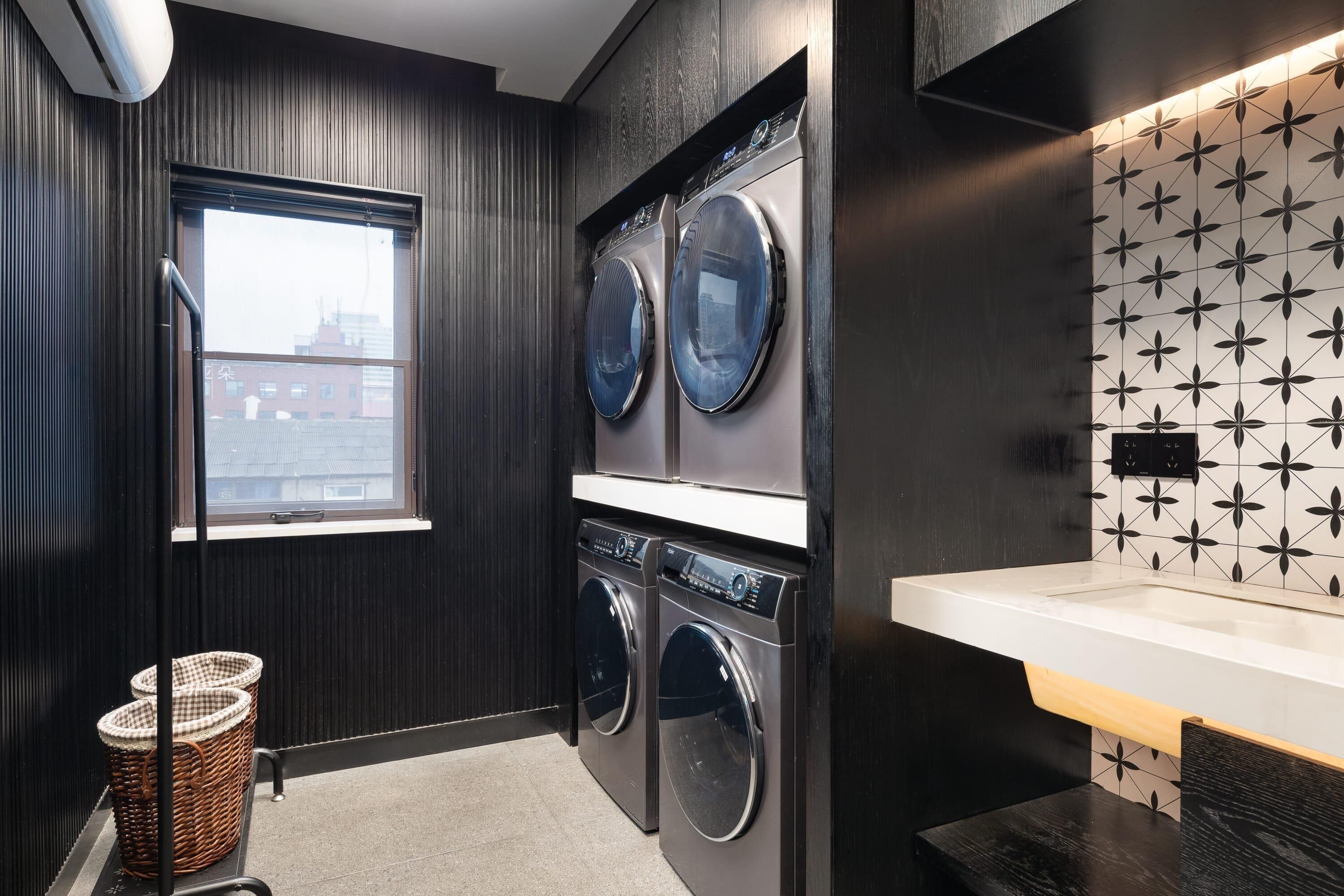 A laundry room with two front-loading washing machines, a window, a wall-mounted sink, and a patterned backsplash.