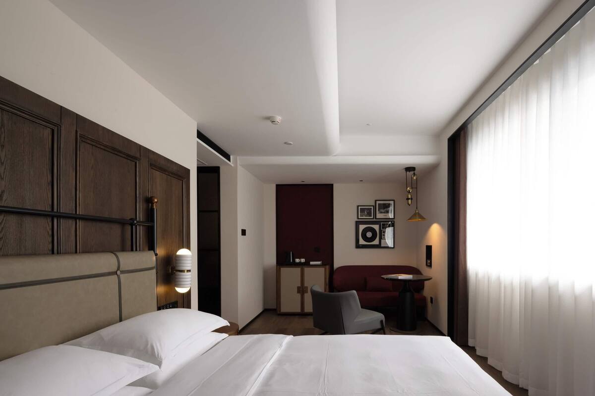 A hotel room with a large bed, a desk, a chair, and a small table. There is a wardrobe with a mirror and a wall-mounted light fixture.