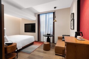 Ein Hotelzimmer mit Bett, Schreibtisch, Stuhl, kleinem Tisch, Fernseher und einem Fenster mit Vorhängen.