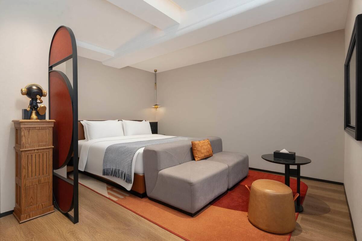 A modern hotel room with a bed, a small sofa, a round ottoman, a side table, and a decorative mirror.