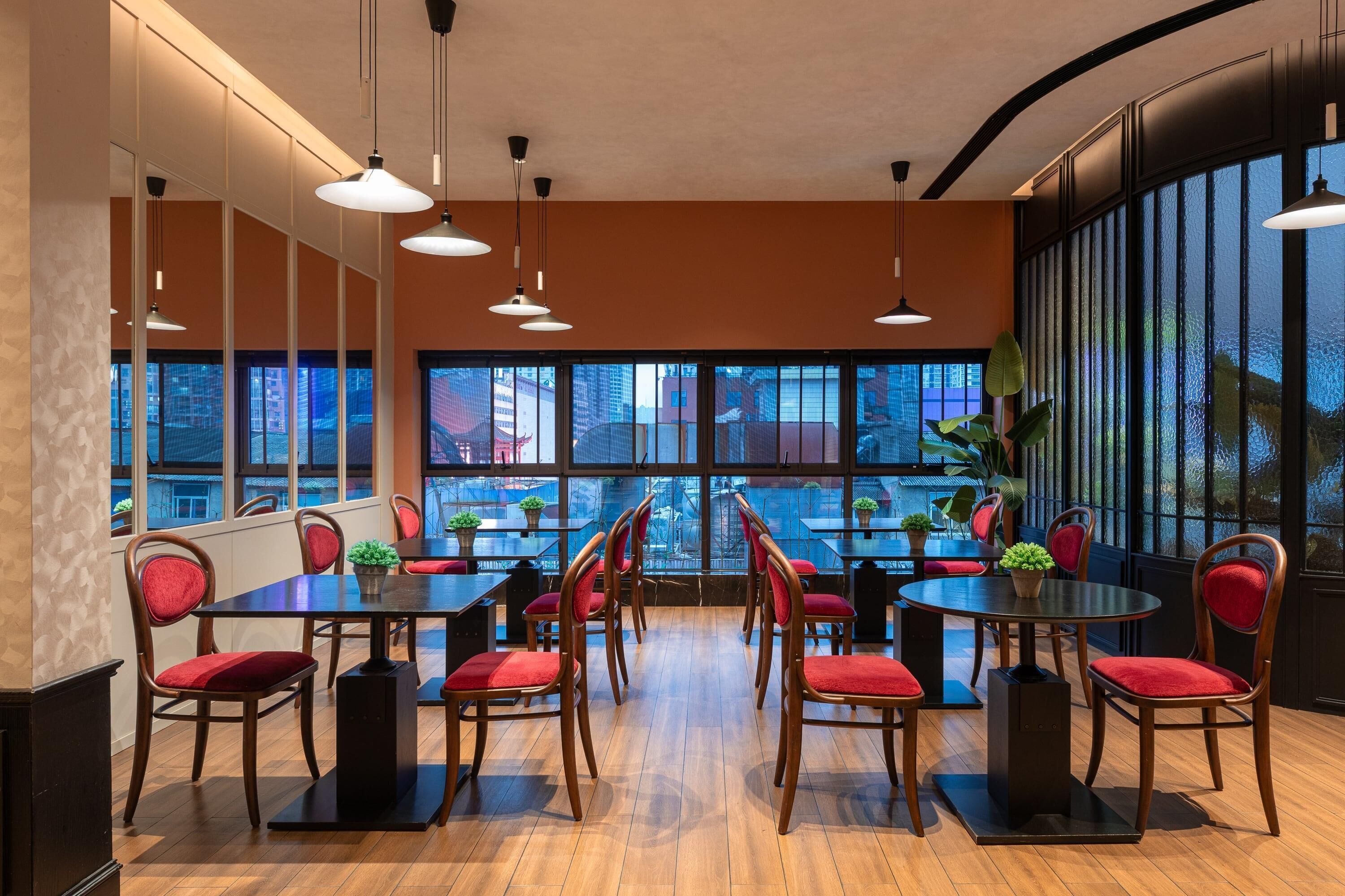 A modern dining area with wooden tables and red chairs, large windows with city views, and pendant lighting.