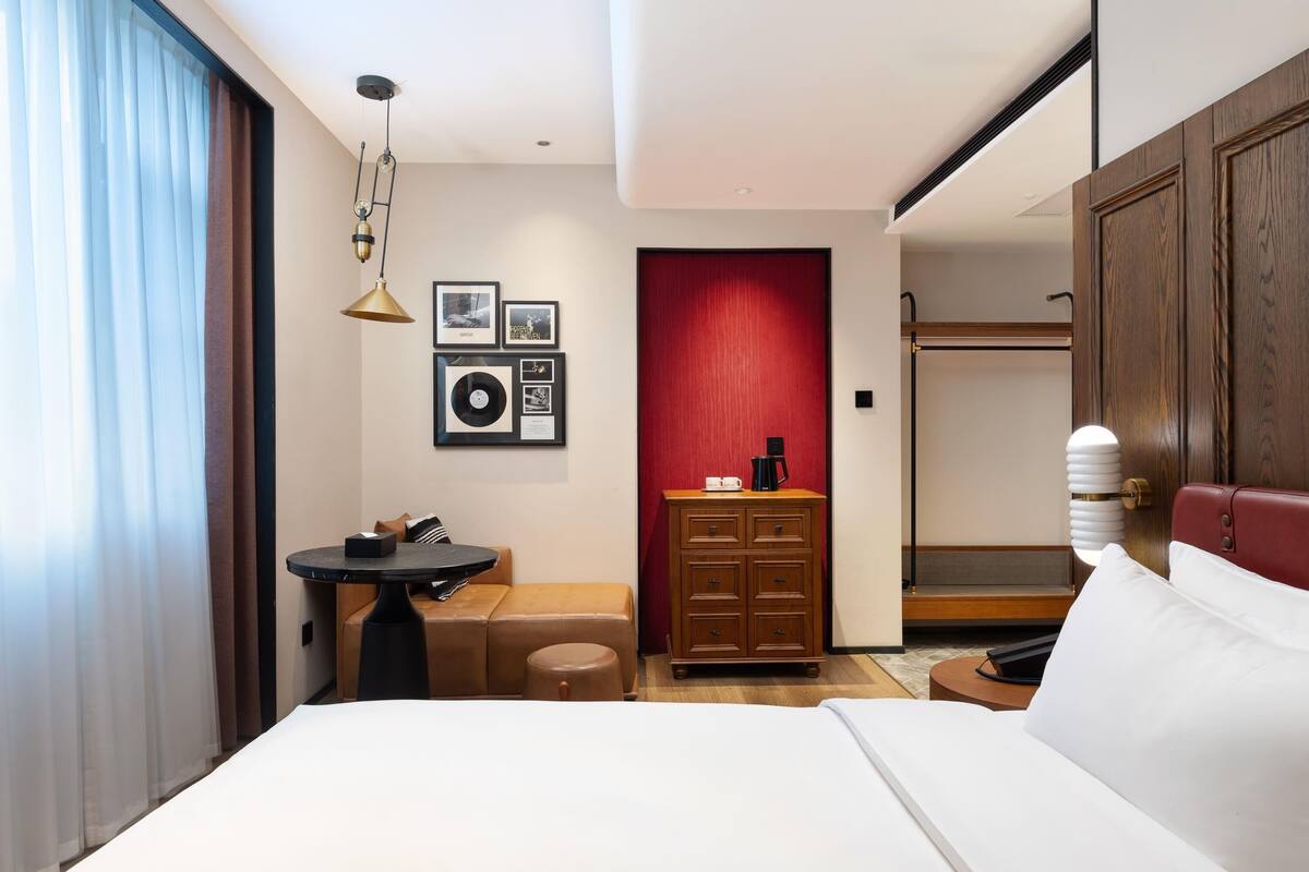 A hotel room with a bed, a small table, a dresser, and a red accent wall.
