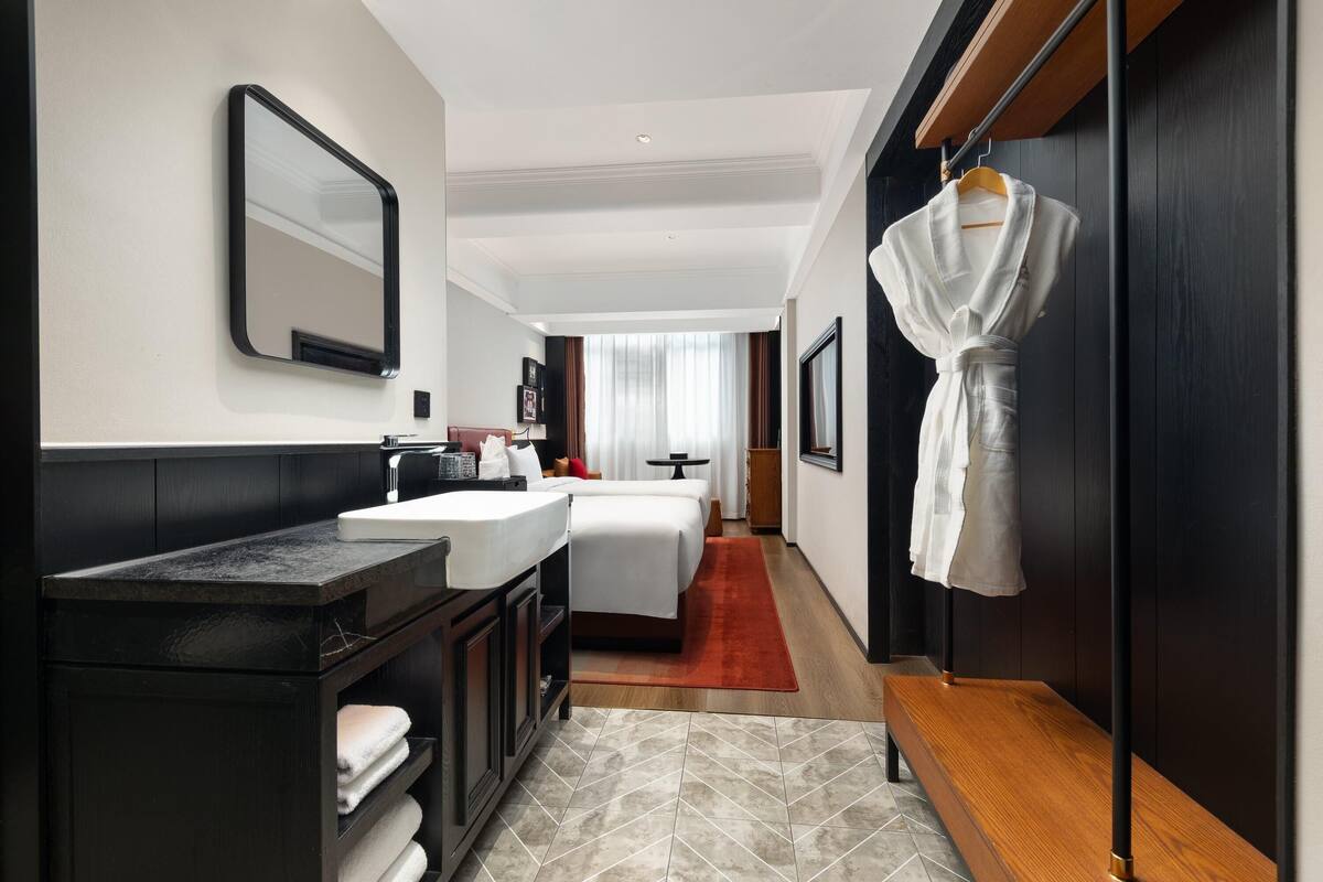 A modern hotel room with a large mirror, a bed, a vanity with a sink, and a robe hanging on the wall.