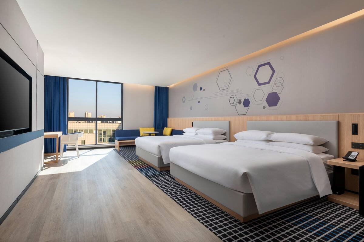 Ein modernes Hotelzimmer mit zwei Betten, einem Schreibtisch, einem Sessel, einem großen Fenster mit Blick auf die Stadt und einem geometrischen Wanddesign.
