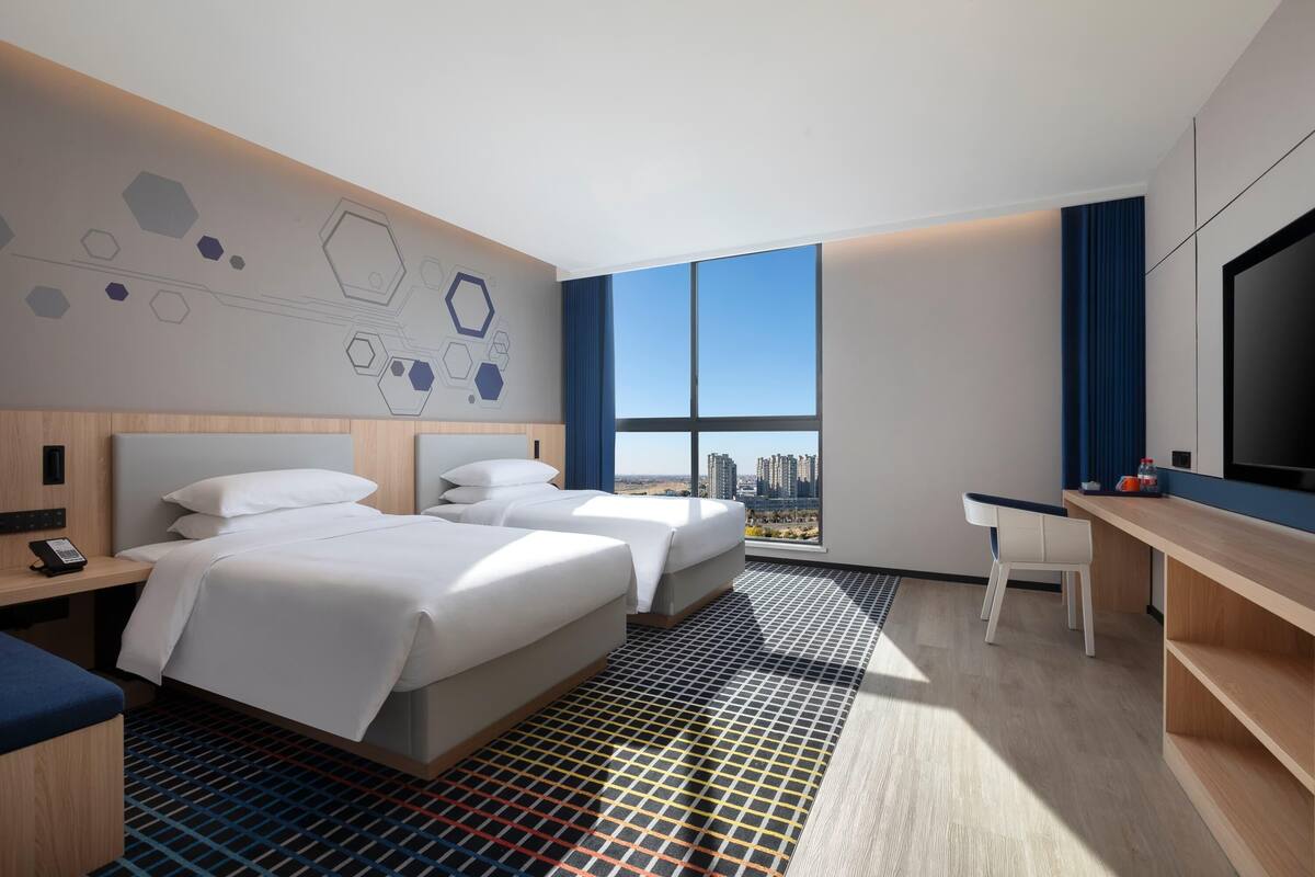 Ein Hotelzimmer mit zwei Betten, einem Schreibtisch, einem Fernseher und einem großen Fenster mit Blick auf die Stadt.
