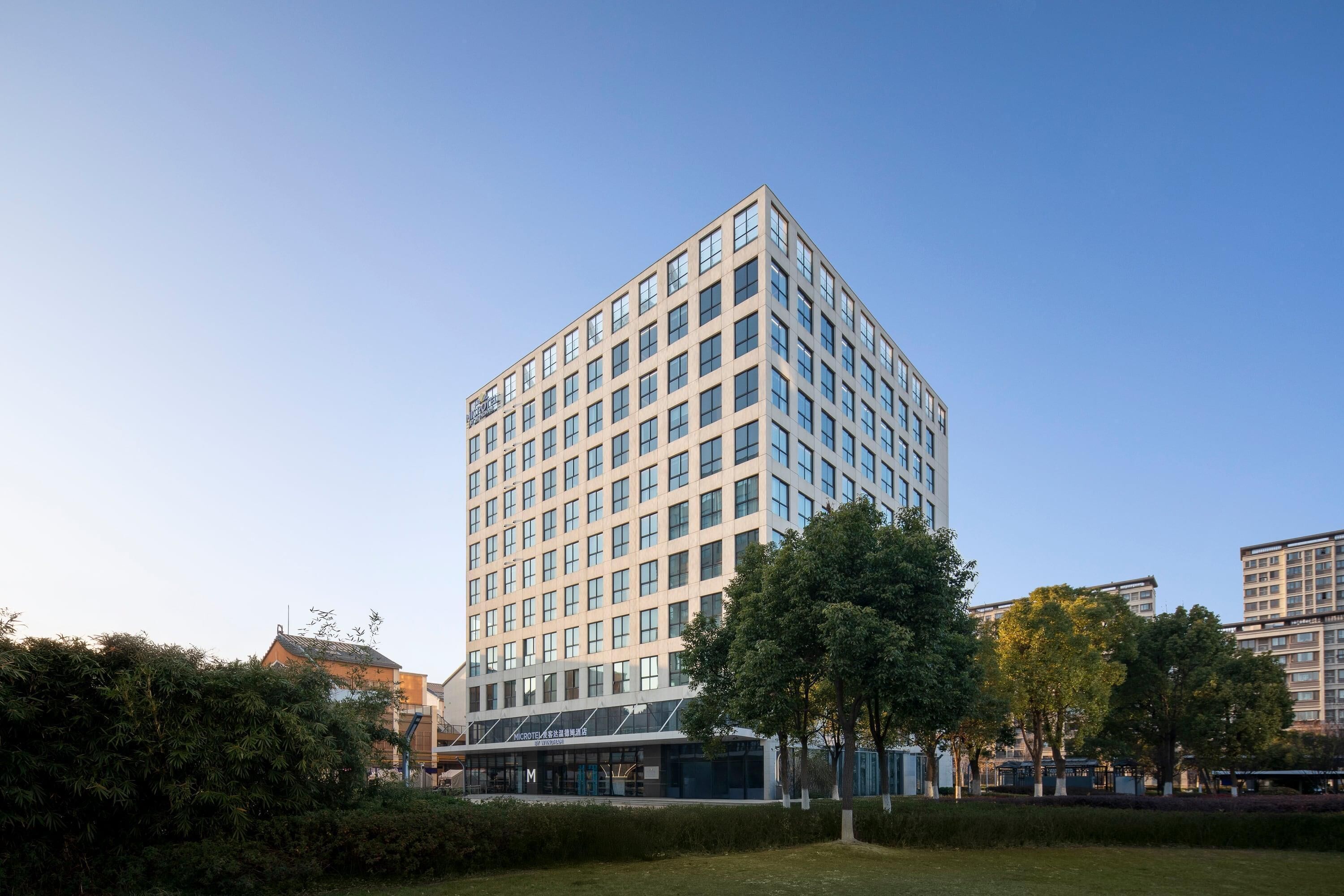 A modern high-rise building with a grid of windows, surrounded by greenery and other buildings.
